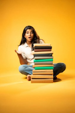 Kitap Yığını 'yla hüzünle oturan, Gergin Hintli Genç Kız Sarı Kusursuz Arkaplanda Bunalmış, Çalışmaların ve Etrafındaki Eğitim Kitapları Yığını ile Akademik Baskının Yükünü Hissediyor