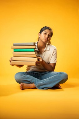 Kitap Yığını 'yla hüzünle oturan, Gergin Hintli Genç Kız Sarı Kusursuz Arkaplanda Bunalmış, Çalışmaların ve Etrafındaki Eğitim Kitapları Yığını ile Akademik Baskının Yükünü Hissediyor