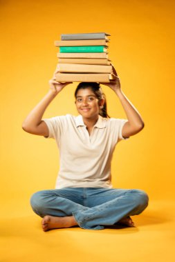 Kitap Yığını 'yla hüzünle oturan, Gergin Hintli Genç Kız Sarı Kusursuz Arkaplanda Bunalmış, Çalışmaların ve Etrafındaki Eğitim Kitapları Yığını ile Akademik Baskının Yükünü Hissediyor