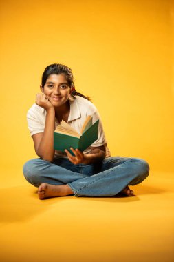Sarı Kusursuz Arkaplanda Otururken Gülümseyerek Bir Kitap Okuyan Kız, Hintli Asyalı Liseli Kız Güvenle Kameraya Bakıyor, Odaklanma, Meraklanma ve Ödev ve Ödev Zamanı Sırasındaki Neşe Gösteriyor