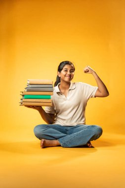 Kitap Yığını Tutarken, Hintli Asyalı Genç Kız Sarı Kusursuz Arkaplanda Mutlulukla Eğitim Kitapları Yığını Göstererek, Parlak ve Neşeli İfadeyle Öğrenme ve Çalışmayı Destekliyor