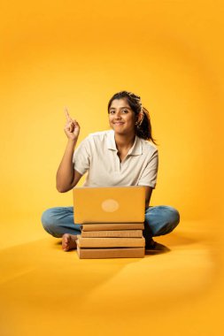 Laptop ve Pointing kullanarak, Hintli Asyalı Ergen Kız Sarı Kusursuz Arkaplanda Oturarak Kitap ve Bilgisayar ile, Dijital Öğrenme, Eğitim Teknolojisi, Çevrimiçi Çalışma Konseptini Destekliyor