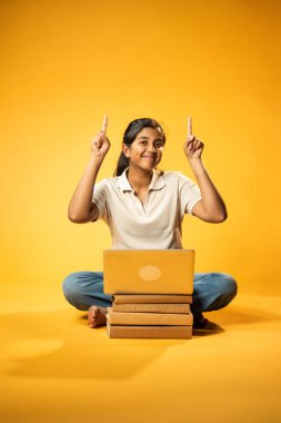 Laptop ve Pointing kullanarak, Hintli Asyalı Ergen Kız Sarı Kusursuz Arkaplanda Oturarak Kitap ve Bilgisayar ile, Dijital Öğrenme, Eğitim Teknolojisi, Çevrimiçi Çalışma Konseptini Destekliyor