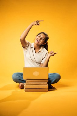 Laptop ve Pointing kullanarak, Hintli Asyalı Ergen Kız Sarı Kusursuz Arkaplanda Oturarak Kitap ve Bilgisayar ile, Dijital Öğrenme, Eğitim Teknolojisi, Çevrimiçi Çalışma Konseptini Destekliyor