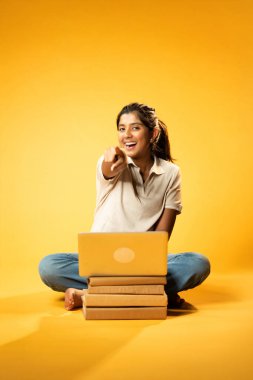 Laptop ve Pointing kullanarak, Hintli Asyalı Ergen Kız Sarı Kusursuz Arkaplanda Oturarak Kitap ve Bilgisayar ile, Dijital Öğrenme, Eğitim Teknolojisi, Çevrimiçi Çalışma Konseptini Destekliyor