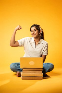 Laptop ve Pointing kullanarak, Hintli Asyalı Genç Kız Sarı Kusursuz Arkaplan Karşısında Oturarak Kitap ve Bilgisayar ile Akademik Başarı Kutlaması ve Dijital Öğrenme Konseptini Destekliyor