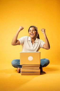 Laptop ve Pointing kullanarak, Hintli Asyalı Genç Kız Sarı Kusursuz Arkaplan Karşısında Oturarak Kitap ve Bilgisayar ile Akademik Başarı Kutlaması ve Dijital Öğrenme Konseptini Destekliyor