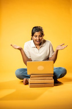 Laptop ve Pointing kullanarak, Hintli Asyalı Genç Kız Sarı Kusursuz Arkaplan Karşısında Oturarak Kitap ve Bilgisayar ile Akademik Başarı Kutlaması ve Dijital Öğrenme Konseptini Destekliyor