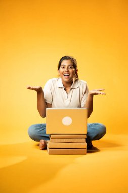 Laptop ve Pointing kullanarak, Hintli Asyalı Genç Kız Sarı Kusursuz Arkaplan Karşısında Oturarak Kitap ve Bilgisayar ile Akademik Başarı Kutlaması ve Dijital Öğrenme Konseptini Destekliyor