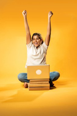 Laptop ve Pointing kullanarak, Hintli Asyalı Genç Kız Sarı Kusursuz Arkaplan Karşısında Oturarak Kitap ve Bilgisayar ile Akademik Başarı Kutlaması ve Dijital Öğrenme Konseptini Destekliyor