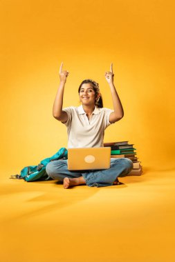 Laptop ve Pointing kullanarak, Hintli Asyalı Genç Kız Sarı Kusursuz Arkaplan Karşısında Oturarak Kitap ve Bilgisayar ile Akademik Başarı Kutlaması ve Dijital Öğrenme Konseptini Destekliyor