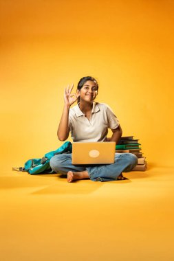 Laptop ve Pointing kullanarak, Hintli Asyalı Genç Kız Sarı Kusursuz Arkaplan Karşısında Oturarak Kitap ve Bilgisayar ile Akademik Başarı Kutlaması ve Dijital Öğrenme Konseptini Destekliyor