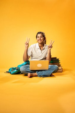 Laptop ve Pointing kullanarak, Hintli Asyalı Genç Kız Sarı Kusursuz Arkaplan Karşısında Oturarak Kitap ve Bilgisayar ile Akademik Başarı Kutlaması ve Dijital Öğrenme Konseptini Destekliyor