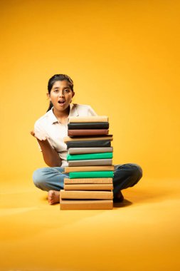 Kitap Yığını 'yla hüzünle oturan, Gergin Hintli Genç Kız Sarı Kusursuz Arkaplanda Bunalmış, Çalışmaların ve Etrafındaki Eğitim Kitapları Yığını ile Akademik Baskının Yükünü Hissediyor