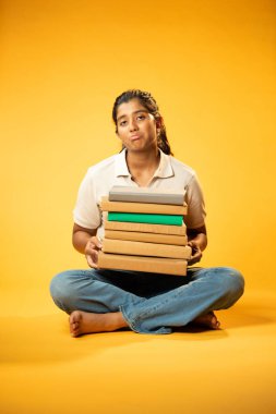 Kitap Yığını 'yla hüzünle oturan, Gergin Hintli Genç Kız Sarı Kusursuz Arkaplanda Bunalmış, Çalışmaların ve Etrafındaki Eğitim Kitapları Yığını ile Akademik Baskının Yükünü Hissediyor