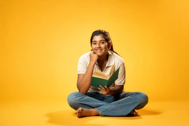 Sarı Kusursuz Arkaplanda Otururken Gülümseyerek Bir Kitap Okuyan Kız, Hintli Asyalı Liseli Kız Güvenle Kameraya Bakıyor, Odaklanma, Meraklanma ve Ödev ve Ödev Zamanı Sırasındaki Neşe Gösteriyor