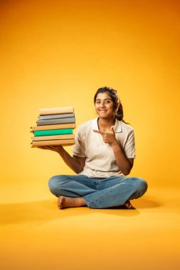 Kitap Yığını Tutarken, Hintli Asyalı Genç Kız Sarı Kusursuz Arkaplanda Mutlulukla Eğitim Kitapları Yığını Göstererek, Parlak ve Neşeli İfadeyle Öğrenme ve Çalışmayı Destekliyor