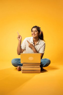 Laptop ve Pointing kullanarak, Hintli Asyalı Ergen Kız Sarı Kusursuz Arkaplanda Oturarak Kitap ve Bilgisayar ile, Dijital Öğrenme, Eğitim Teknolojisi, Çevrimiçi Çalışma Konseptini Destekliyor