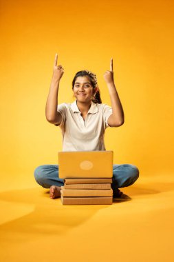 Laptop ve Pointing kullanarak, Hintli Asyalı Ergen Kız Sarı Kusursuz Arkaplanda Oturarak Kitap ve Bilgisayar ile, Dijital Öğrenme, Eğitim Teknolojisi, Çevrimiçi Çalışma Konseptini Destekliyor