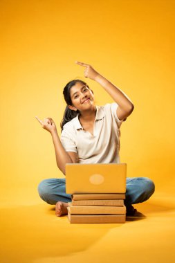 Laptop ve Pointing kullanarak, Hintli Asyalı Ergen Kız Sarı Kusursuz Arkaplanda Oturarak Kitap ve Bilgisayar ile, Dijital Öğrenme, Eğitim Teknolojisi, Çevrimiçi Çalışma Konseptini Destekliyor