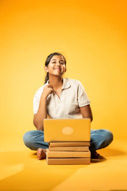 Laptop ve Pointing kullanarak, Hintli Asyalı Genç Kız Sarı Kusursuz Arkaplan Karşısında Oturarak Kitap ve Bilgisayar ile Akademik Başarı Kutlaması ve Dijital Öğrenme Konseptini Destekliyor