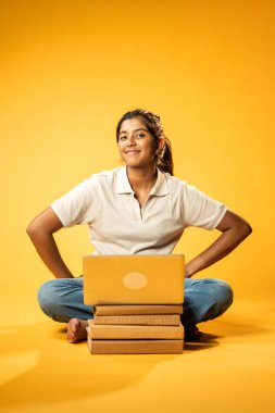 Laptop ve Pointing kullanarak, Hintli Asyalı Genç Kız Sarı Kusursuz Arkaplan Karşısında Oturarak Kitap ve Bilgisayar ile Akademik Başarı Kutlaması ve Dijital Öğrenme Konseptini Destekliyor