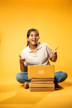 Laptop ve Pointing kullanarak, Hintli Asyalı Genç Kız Sarı Kusursuz Arkaplan Karşısında Oturarak Kitap ve Bilgisayar ile Akademik Başarı Kutlaması ve Dijital Öğrenme Konseptini Destekliyor