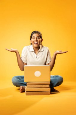 Laptop ve Pointing kullanarak, Hintli Asyalı Genç Kız Sarı Kusursuz Arkaplan Karşısında Oturarak Kitap ve Bilgisayar ile Akademik Başarı Kutlaması ve Dijital Öğrenme Konseptini Destekliyor