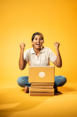 Laptop ve Pointing kullanarak, Hintli Asyalı Genç Kız Sarı Kusursuz Arkaplan Karşısında Oturarak Kitap ve Bilgisayar ile Akademik Başarı Kutlaması ve Dijital Öğrenme Konseptini Destekliyor