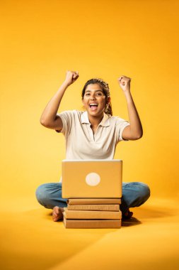 Laptop ve Pointing kullanarak, Hintli Asyalı Genç Kız Sarı Kusursuz Arkaplan Karşısında Oturarak Kitap ve Bilgisayar ile Akademik Başarı Kutlaması ve Dijital Öğrenme Konseptini Destekliyor