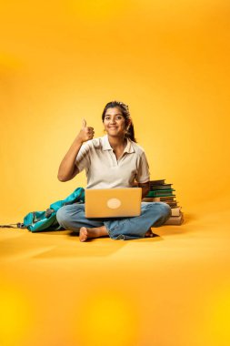 Laptop ve Pointing kullanarak, Hintli Asyalı Genç Kız Sarı Kusursuz Arkaplan Karşısında Oturarak Kitap ve Bilgisayar ile Akademik Başarı Kutlaması ve Dijital Öğrenme Konseptini Destekliyor