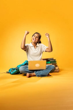 Laptop ve Pointing kullanarak, Hintli Asyalı Genç Kız Sarı Kusursuz Arkaplan Karşısında Oturarak Kitap ve Bilgisayar ile Akademik Başarı Kutlaması ve Dijital Öğrenme Konseptini Destekliyor