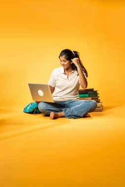 Laptop ve Pointing kullanarak, Hintli Asyalı Genç Kız Sarı Kusursuz Arkaplan Karşısında Oturarak Kitap ve Bilgisayar ile Akademik Başarı Kutlaması ve Dijital Öğrenme Konseptini Destekliyor