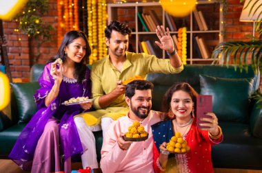 Geleneksel şenlikte Hintli Asyalı genç çiftler Diwali 'ye sesleniyor. Deepavali' yi kutlamak için dekore edilmiş evde laddu barfi tatlıları tutuyor ve akıllı telefon görüşmelerine katılıyorlar.