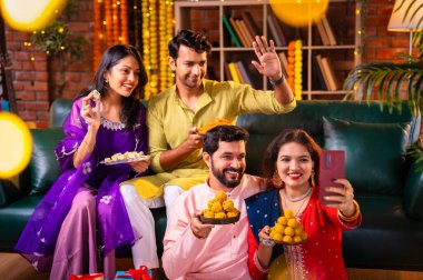 Geleneksel şenlikte Hintli Asyalı genç çiftler Diwali 'ye sesleniyor. Deepavali' yi kutlamak için dekore edilmiş evde laddu barfi tatlıları tutuyor ve akıllı telefon görüşmelerine katılıyorlar.