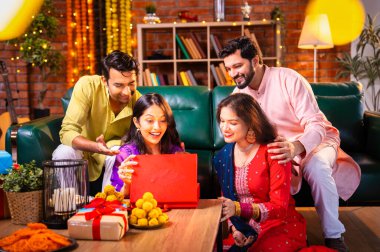 Geleneksel şenlik kıyafetleri içindeki Hintli Hindu çift, bir arkadaştan Diwali hediyesi alarak sürpriz bir şekilde arkadaşlarının hediyelerini açıyor ve güzel dekore edilmiş evlerinde neşeyle sarılıyorlar.