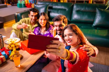 Arkadaşlarıyla Diwali selfie çeken Hindu genç evli çift Hindu festivali Deepavali 'yi kutlamak için dekore edilmiş evde laddu barfi şekerlerini tıklayarak selfie çekiyor.