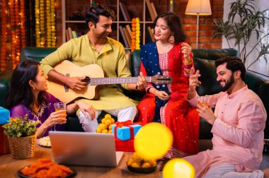 Diwali kutlamaları sırasında şarkı söyleyen arkadaşlar, Hint Asyalı genç evli çift geleneksel şenlik kıyafetleriyle gitar ve müzik eşliğinde ev sahipliği yapıyor dekore edilmiş evlerde Deepavali gecesinin tadını çıkarıyorlar.