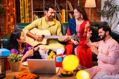 Diwali kutlamaları sırasında şarkı söyleyen arkadaşlar, Hint Asyalı genç evli çift geleneksel şenlik kıyafetleriyle gitar ve müzik eşliğinde ev sahipliği yapıyor dekore edilmiş evlerde Deepavali gecesinin tadını çıkarıyorlar.