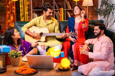 Diwali kutlamaları sırasında şarkı söyleyen arkadaşlar, Hint Asyalı genç evli çift geleneksel şenlik kıyafetleriyle gitar ve müzik eşliğinde ev sahipliği yapıyor dekore edilmiş evlerde Deepavali gecesinin tadını çıkarıyorlar.