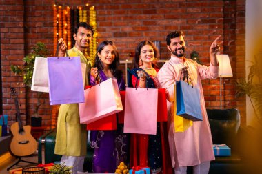 Gösterişli Diwali alışveriş torbaları ve tebessümlerle selamlaşan Hintli Asyalı genç evli çiftler dekore edilmiş evlerin iç kısımlarında geleneksel şenlik kıyafetleri içinde ve kameraya neşe dolu alışveriş torbaları gösteriyorlar.