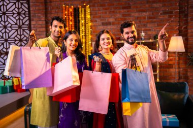 Gösterişli Diwali alışveriş torbaları ve tebessümlerle selamlaşan Hintli Asyalı genç evli çiftler dekore edilmiş evlerin iç kısımlarında geleneksel şenlik kıyafetleri içinde ve kameraya neşe dolu alışveriş torbaları gösteriyorlar.