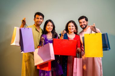 Gösterişli Diwali alışveriş torbaları ve tebessümlerle selamlaşan Hintli Asyalı genç evli çiftler dekore edilmiş evlerin iç kısımlarında geleneksel şenlik kıyafetleri içinde ve kameraya neşe dolu alışveriş torbaları gösteriyorlar.