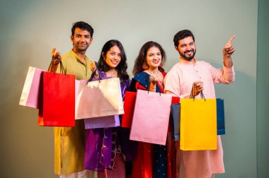 Gösterişli Diwali alışveriş torbaları ve tebessümlerle selamlaşan Hintli Asyalı genç evli çiftler dekore edilmiş evlerin iç kısımlarında geleneksel şenlik kıyafetleri içinde ve kameraya neşe dolu alışveriş torbaları gösteriyorlar.