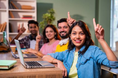 Üniversite öğrencileri, bilgisayar, kitap ve dostça üniversite kütüphanesi ortamındaki notlar üzerinde çalışırken boş alanı heyecanlı bir şekilde işaret ederek başarıyı kutluyorlar.