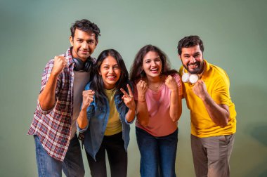 Üniversite öğrencileri zaferlerini kutluyorlar, Hintli üniversite gençleri yeşil duvara karşı ayakta duruyorlar, gülümsüyorlar, tezahürat yapıyorlar, akademik başarı üstüne sevinçle yumruklarını kaldırıyorlar.