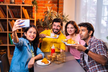 Kafede samosa ve chai içerken selfie çeken Hintli kolej öğrencileri, arkadaşlarıyla birlikte eğlenceli grup fotoğrafları çekerken aynı zamanda kapalı bir restoranda arkadaşlarıyla birlikte akıllı telefon kullanıyorlar.