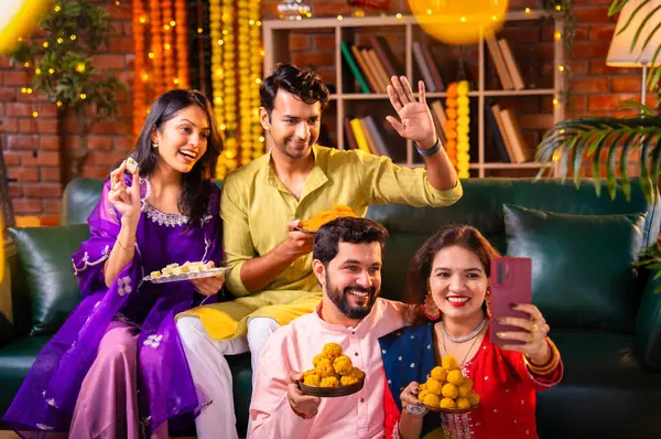 Geleneksel şenlikte Hintli Asyalı genç çiftler Diwali 'ye sesleniyor. Deepavali' yi kutlamak için dekore edilmiş evde laddu barfi tatlıları tutuyor ve akıllı telefon görüşmelerine katılıyorlar.