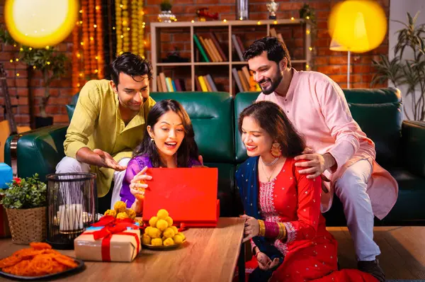 Geleneksel şenlik kıyafetleri içindeki Hintli Hindu çift, bir arkadaştan Diwali hediyesi alarak sürpriz bir şekilde arkadaşlarının hediyelerini açıyor ve güzel dekore edilmiş evlerinde neşeyle sarılıyorlar.