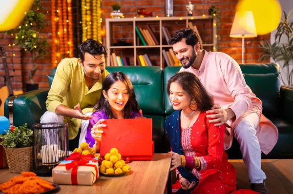Geleneksel şenlik kıyafetleri içindeki Hintli Hindu çift, bir arkadaştan Diwali hediyesi alarak sürpriz bir şekilde arkadaşlarının hediyelerini açıyor ve güzel dekore edilmiş evlerinde neşeyle sarılıyorlar.